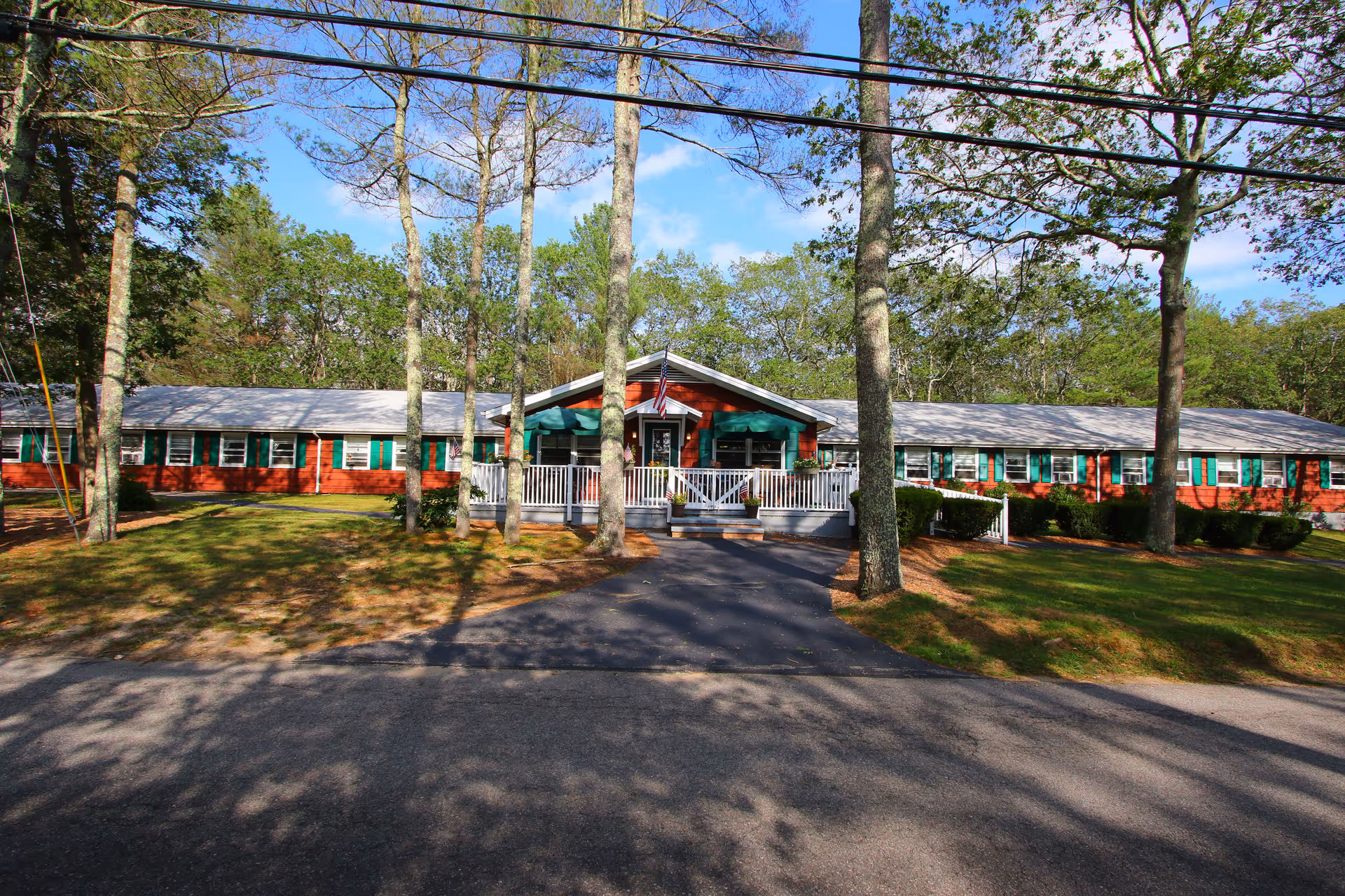 Front exterior view of a single-story red brick building with green window shutters and a white porch railing. The building is surrounded by trees and a grassy lawn under a blue sky with some clouds.