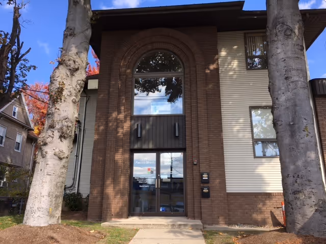 Brick building entrance with an arched window, glass doors, and two large trees flanking the sidewalk.