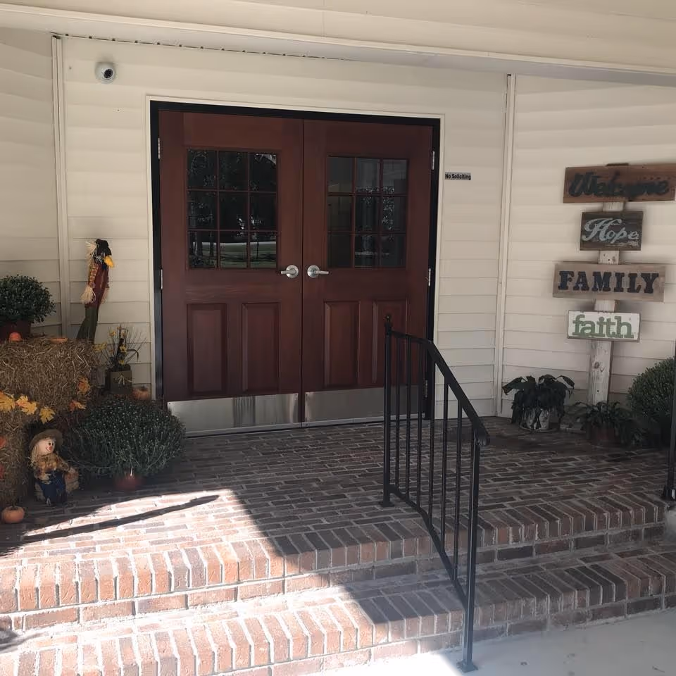 Entrance to a building with double wooden doors featuring glass panes. The entrance has brick steps with a black metal handrail. To the right of the doors, there is a wooden signpost with the words 'Welcome', 'Hope', 'FAMILY', and 'faith'. There are potted plants and autumn-themed decorations including a scarecrow and a small doll on the left side of the entrance.
