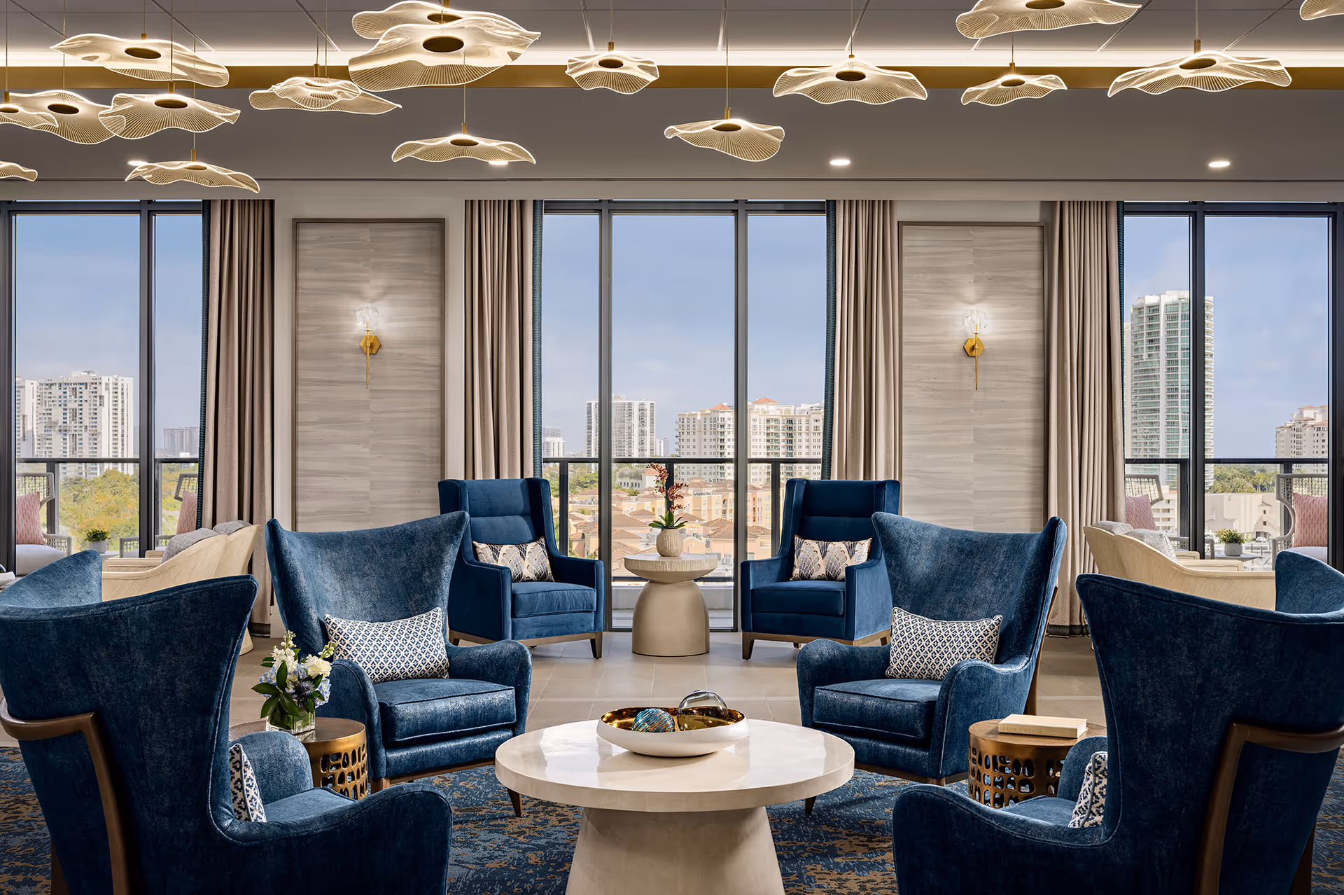 Bright lounge area with blue armchairs, circular table, and expansive city-view windows.