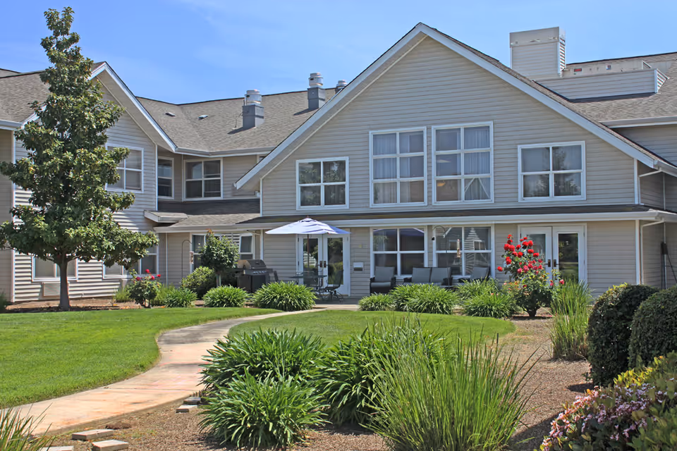 Exterior view of a senior living facility building with beige siding and multiple windows. The building has a well-maintained garden with green bushes, flowering plants, and a tree. A curved concrete pathway leads to a patio area with outdoor furniture and an umbrella. The sky is clear and blue.