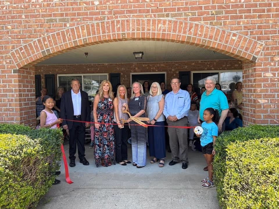 A group of adults and children stand under a brick arch at an entrance holding a red ribbon for a ribbon-cutting ceremony.