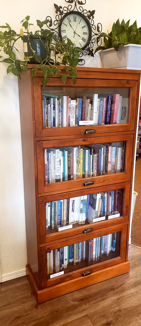 Wooden glass-front bookcase with plants and a clock in a cozy interior.