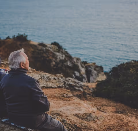 An elderly man sits on a rocky coastal overlook, gazing at the sea.