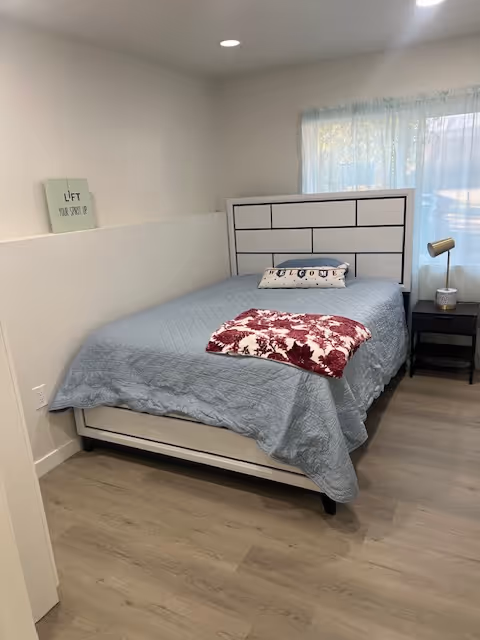 Cozy bedroom in a senior facility featuring a bed, nightstand, lamp, and window.