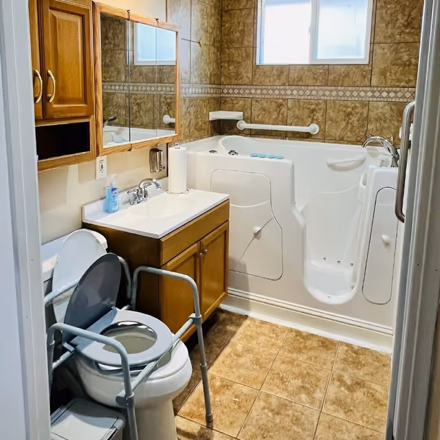 Accessible bathroom featuring sink, mirrored cabinet, toilet with support frame, and walk-in tub with grab bars.