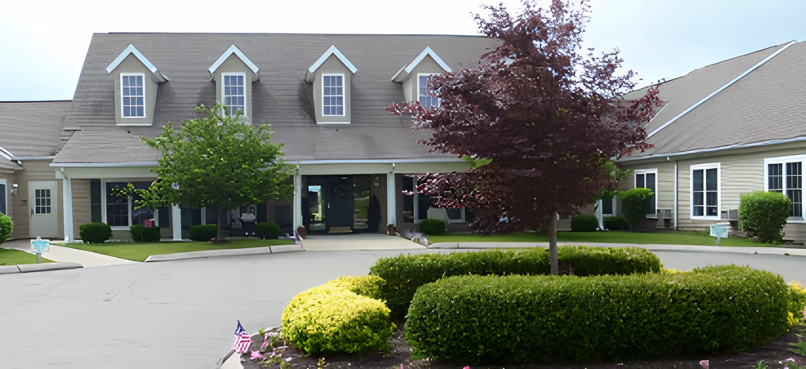 Front entrance of a senior living building with dormer windows, a circular driveway, and landscaped hedges and trees.
