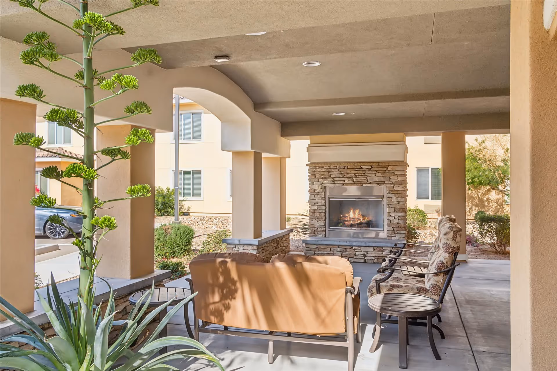 Covered outdoor seating area with cushioned chairs and a loveseat arranged around a stone fireplace with a fire burning. The area is supported by beige columns and overlooks a garden with shrubs and a parked car in the background.