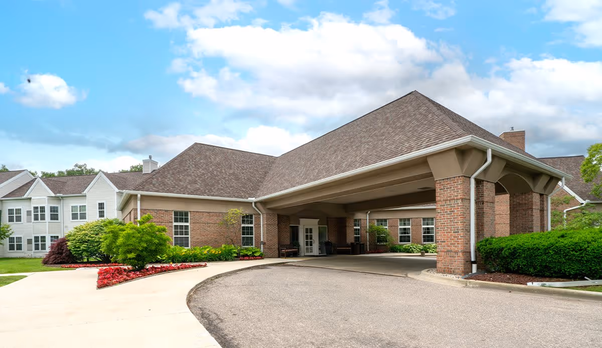 Exterior view of Autumn House Bloomfield Hills showing a brick building with a large covered entrance driveway, surrounded by well-maintained landscaping including green bushes and red flowers under a partly cloudy blue sky.