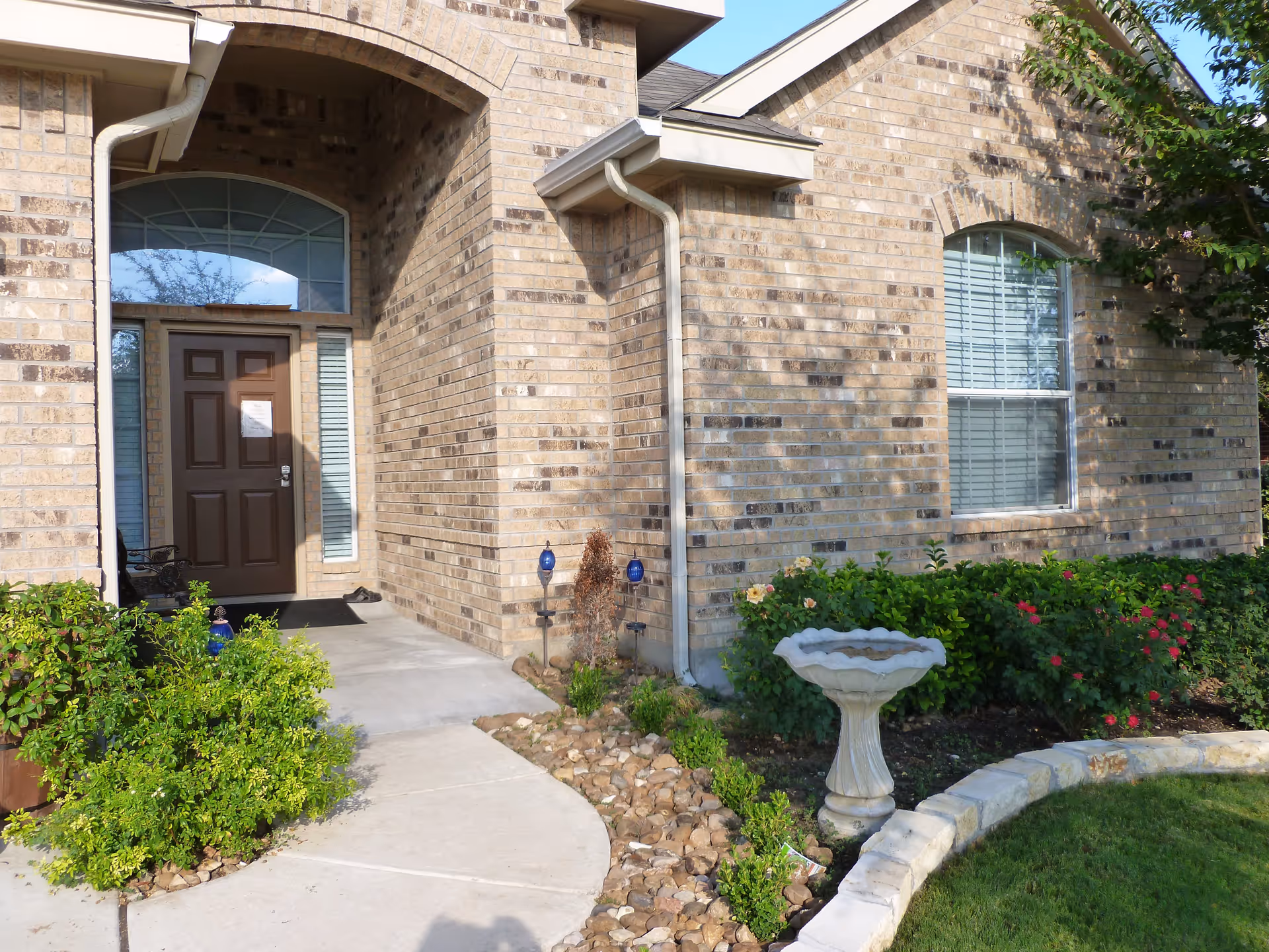 Front entrance of a brick building with a brown door, arched window above the door, and a window with white blinds to the right. There is a concrete pathway leading to the door, surrounded by green bushes, flowering plants, a birdbath, and a small stone border along the grass.