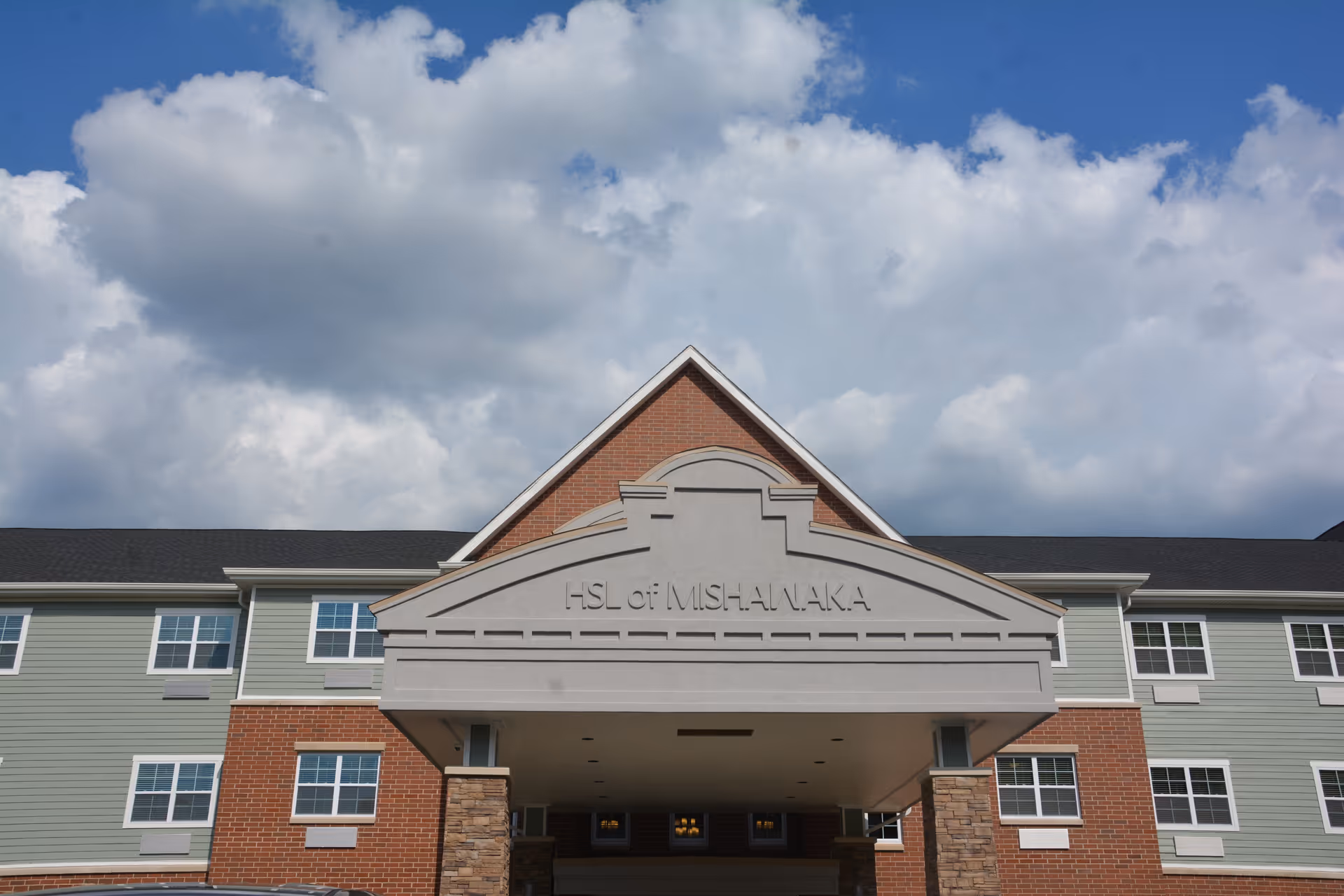 Front exterior view of a senior living facility building with a covered entrance. The building has a combination of brick and light green siding with multiple windows. The sky above is partly cloudy.