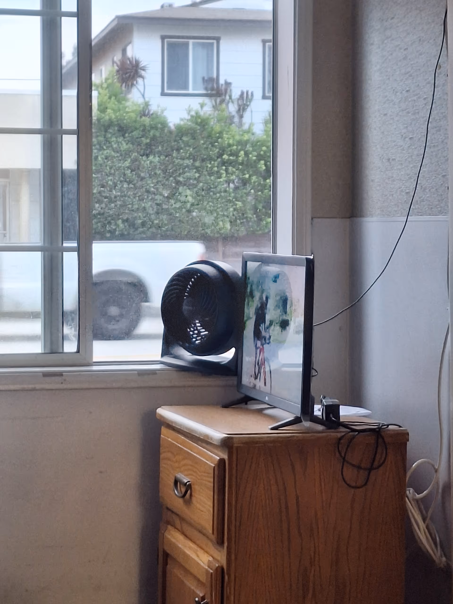A wooden cabinet with a monitor and fan sits beside a window in an indoor room.