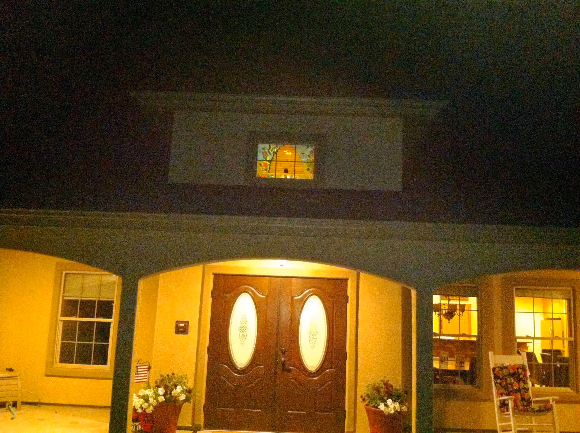 Night view of the front entrance of a building with double wooden doors featuring oval glass panels. The building has a covered porch with two large flower pots on either side of the entrance. There are windows on both sides of the door, and a small stained glass window above the porch. The interior lights are on, illuminating the entrance area.