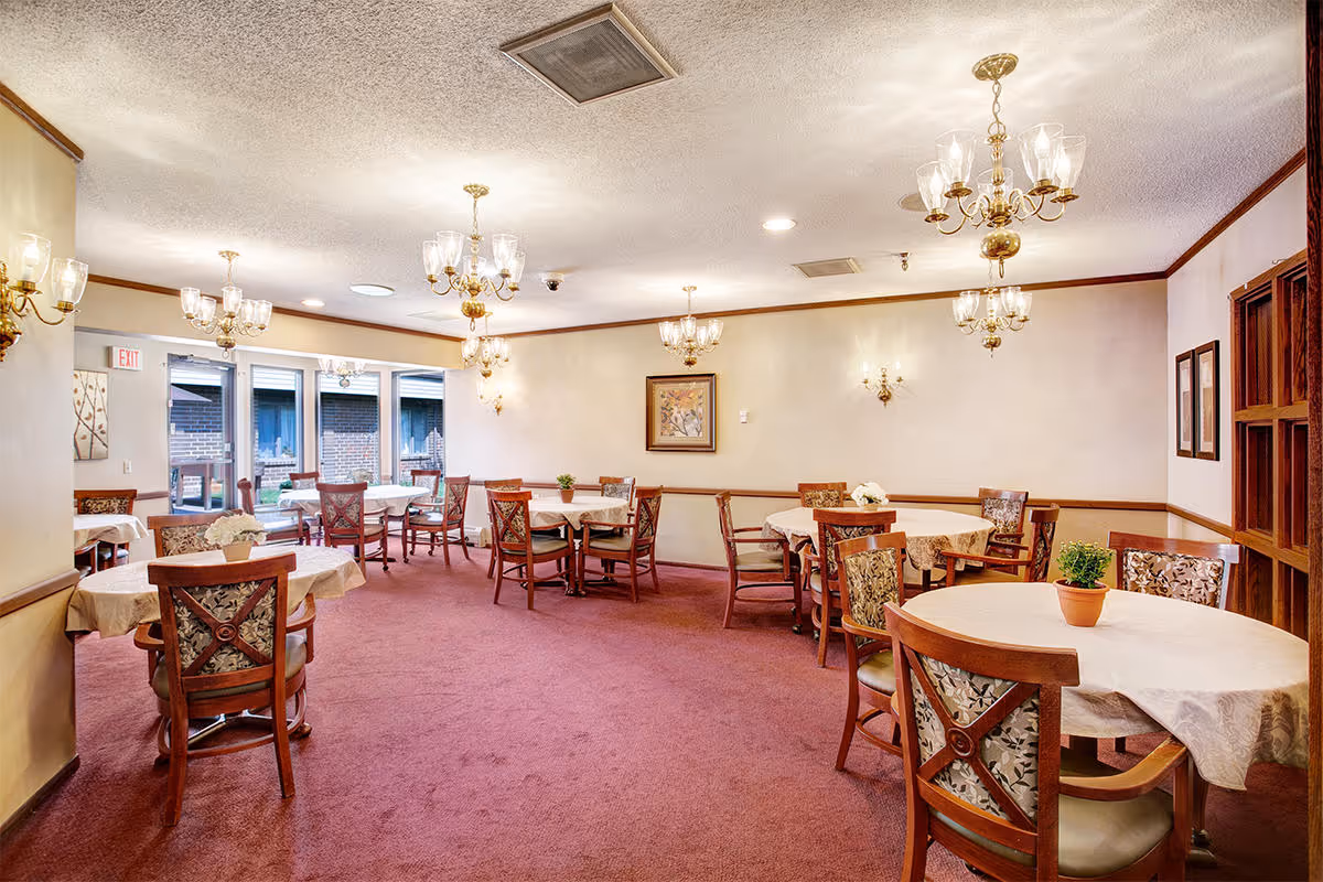 A dining room with multiple round tables covered with white tablecloths, each surrounded by wooden chairs with patterned cushions. The room has a red carpet, cream-colored walls with wooden trim, and several brass chandeliers and wall sconces providing warm lighting. Large windows and a glass door lead to an outdoor area.