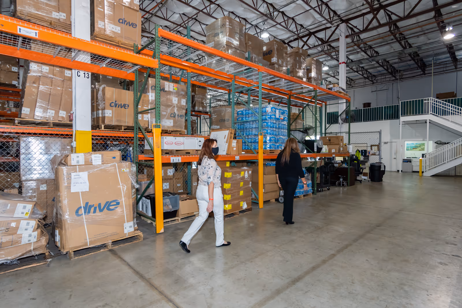 Inside a large warehouse with orange steel shelves, stacked boxes, and two people walking.
