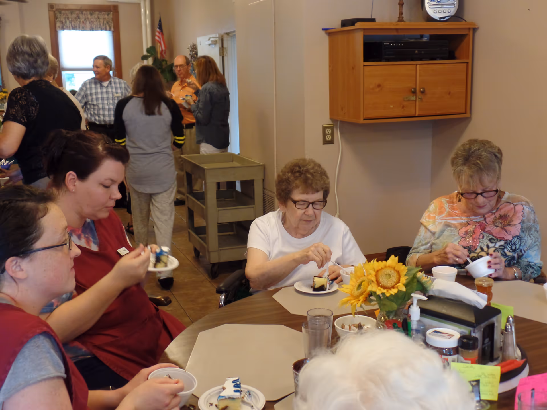 A group of elderly and middle-aged people sitting around a table eating cake and ice cream in a communal dining area. The table has a centerpiece with sunflowers and various condiments. In the background, more people are standing and talking near a window with an American flag beside it.