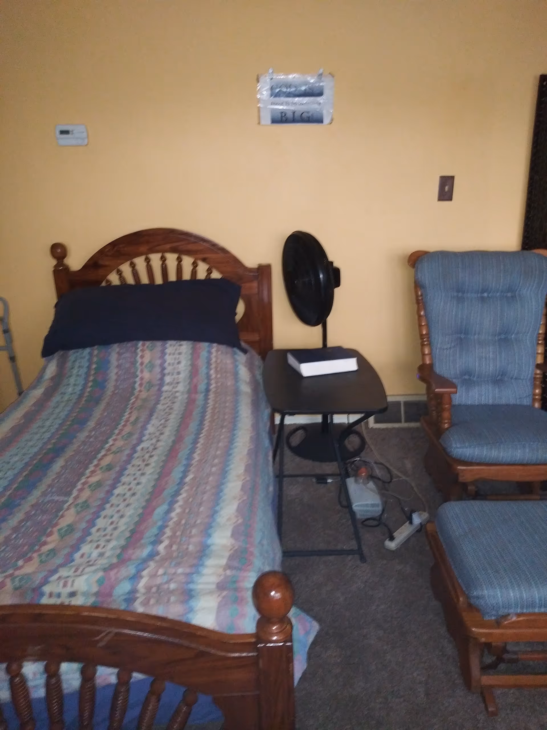 Wooden bed with quilted bedding, side table, fan, and blue upholstered chairs in a small bedroom.