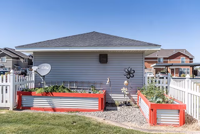 Outdoor garden area with two raised garden beds painted red and silver, surrounded by a white picket fence. A gray building with a sloped roof is in the background, along with a satellite dish and decorative garden ornaments. Other residential buildings are visible further behind.