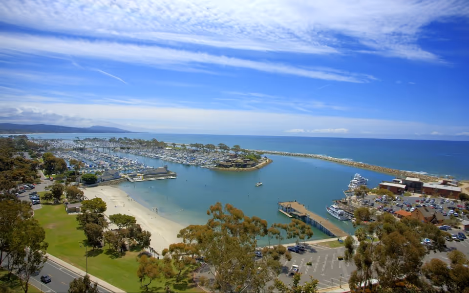 Aerial view of a marina, beach, and harbor with boats and parking.