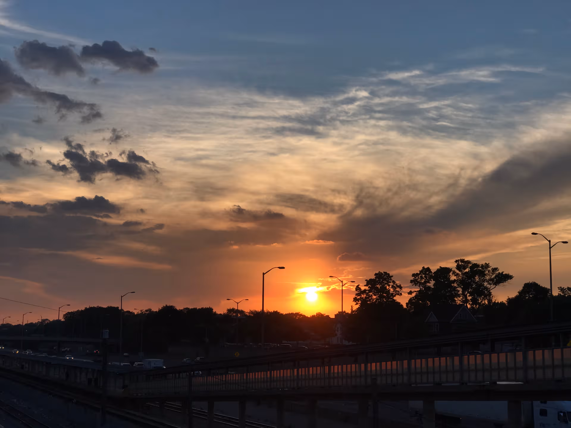 Sunset over a highway with streetlights, silhouetted trees, and a distant horizon.