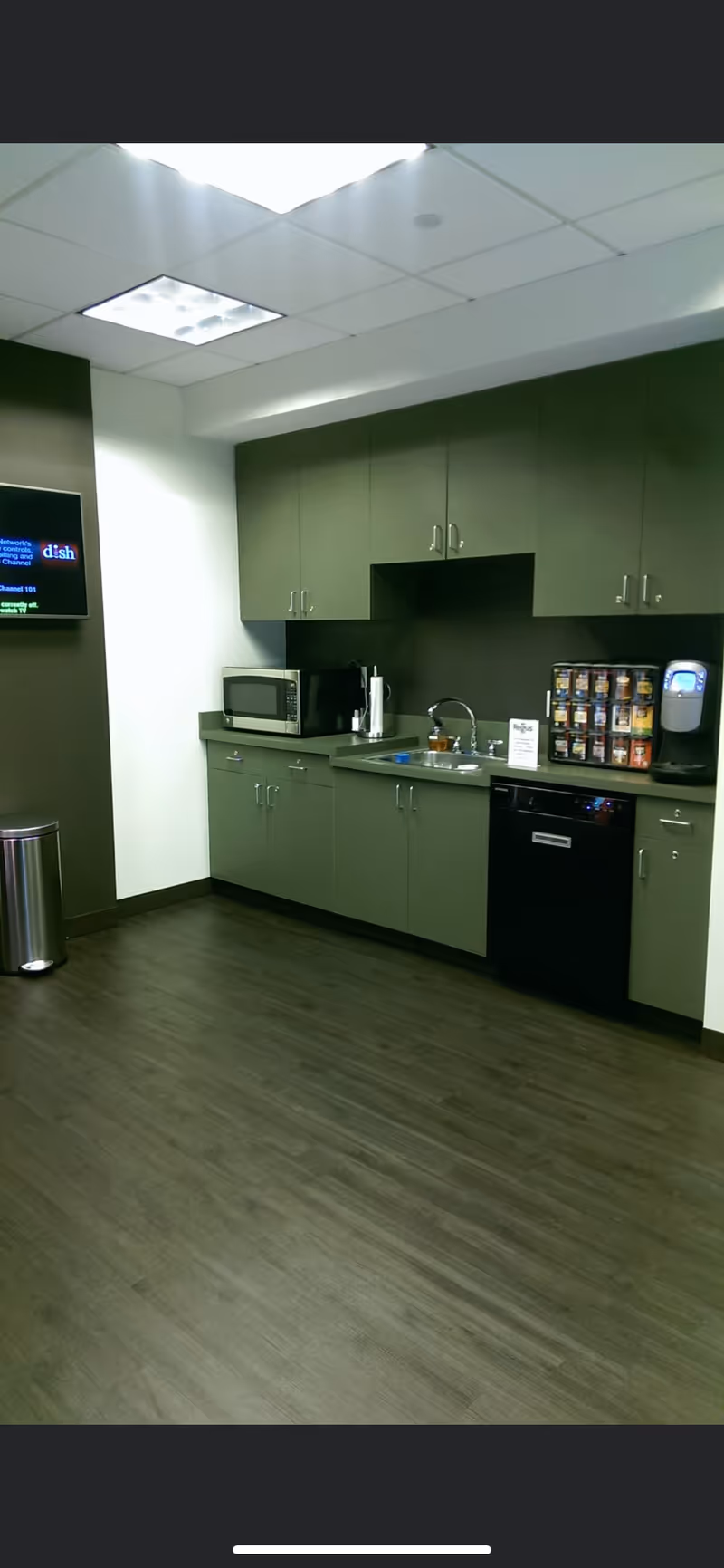 Grey-green kitchenette with cabinets, sink, microwave, coffee maker, and a beverage vending machine.
