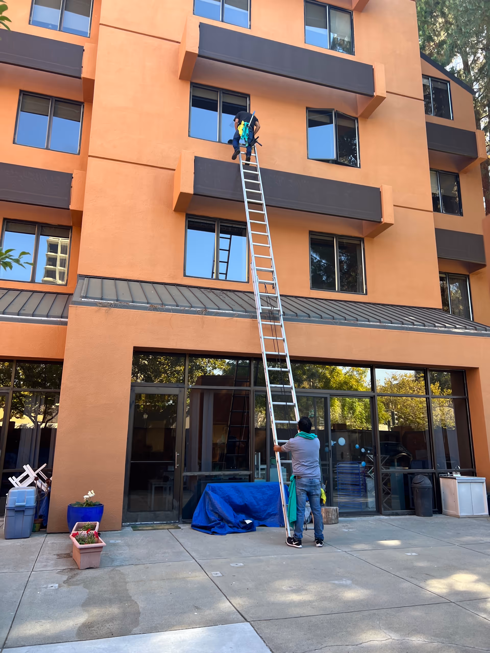 Two workers stabilize a long ladder against an orange multi-story building exterior.