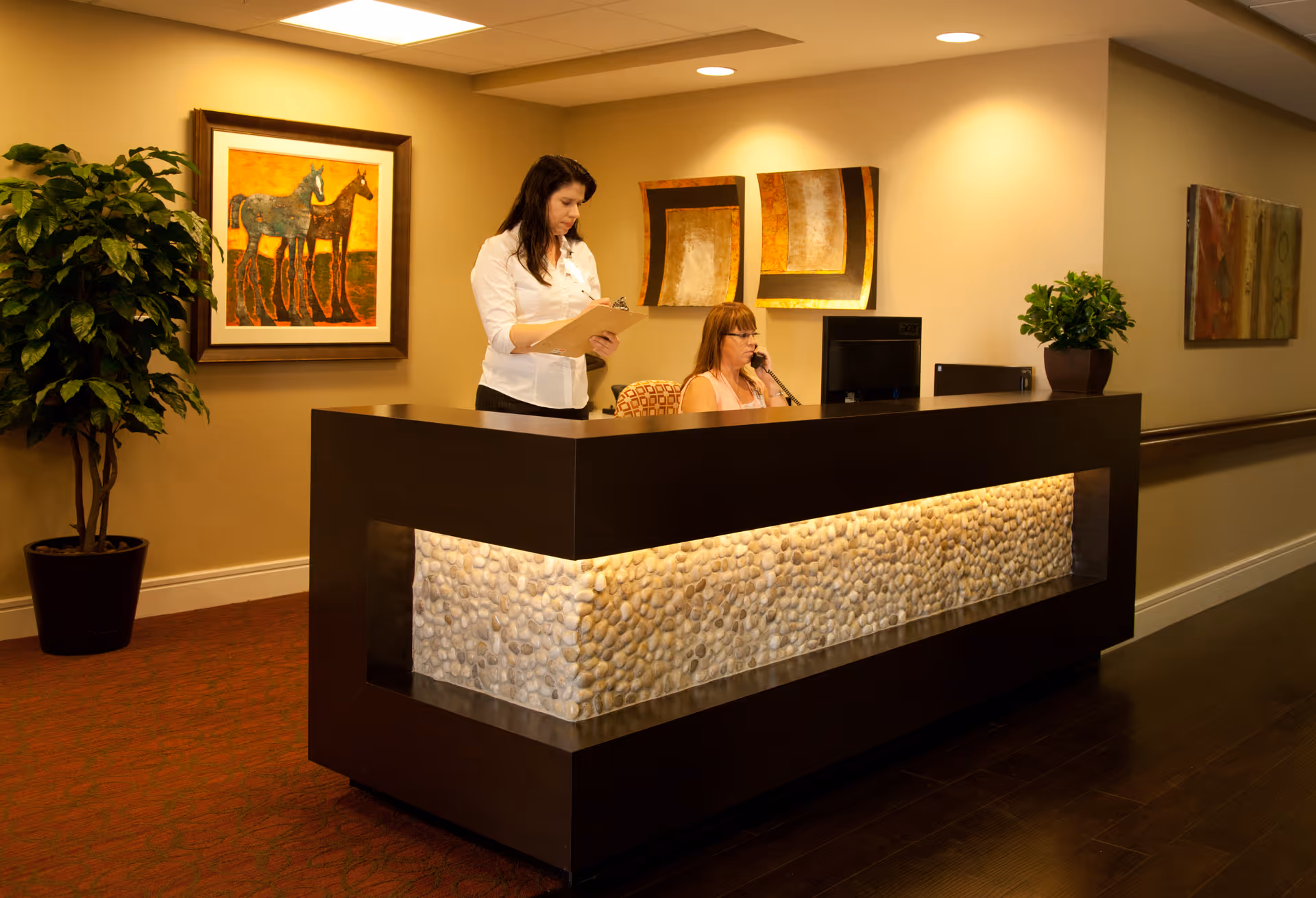 Reception area with a modern dark wood desk featuring a stone panel illuminated from behind. Two women are at the desk, one standing and holding a clipboard, the other seated and speaking on the phone. The walls are decorated with framed artwork and potted plants are placed nearby.