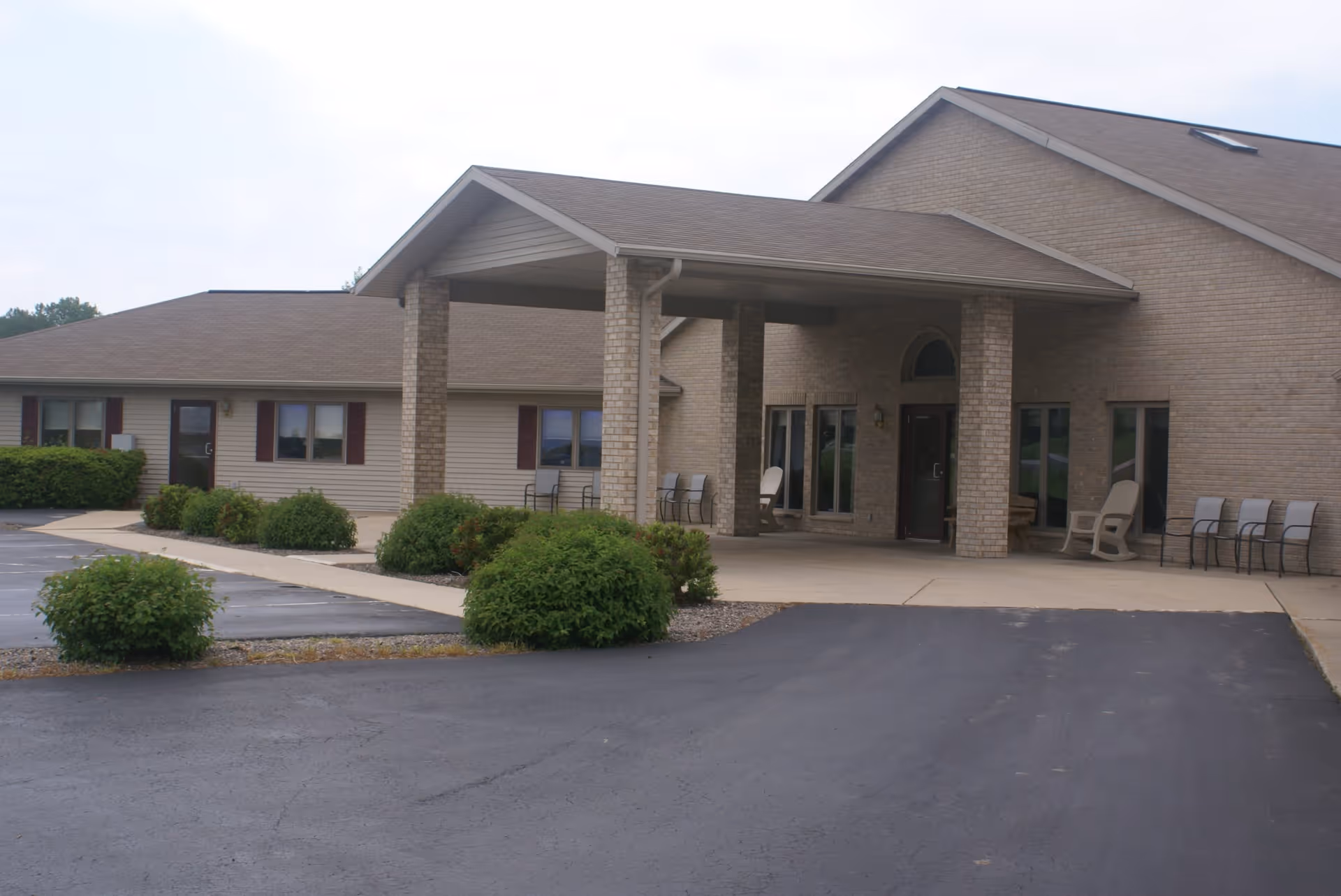 Exterior view of a single-story senior living facility building with a covered entrance supported by brick columns. There are several chairs placed near the entrance and bushes along the walkway and parking area.