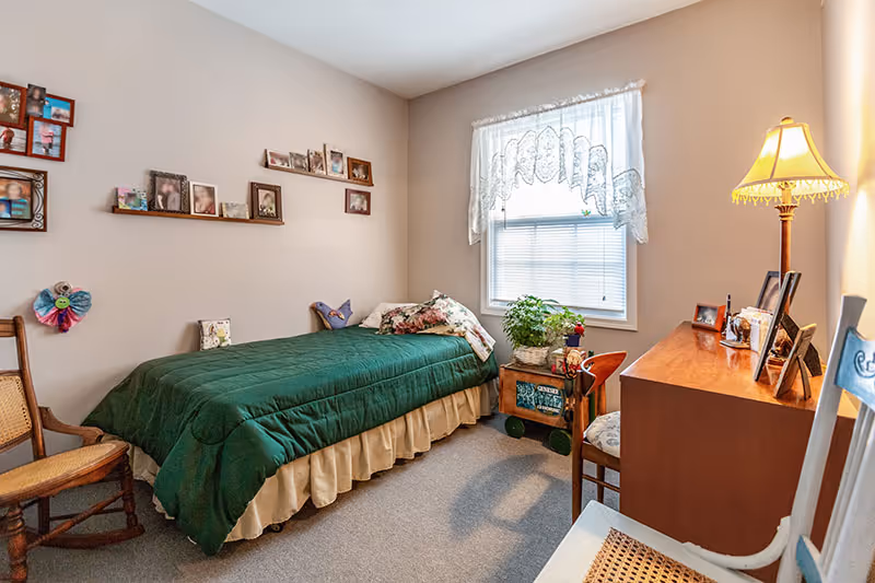 Cozy bedroom with green bedspread, desk, chair, photos on the wall, and a window.