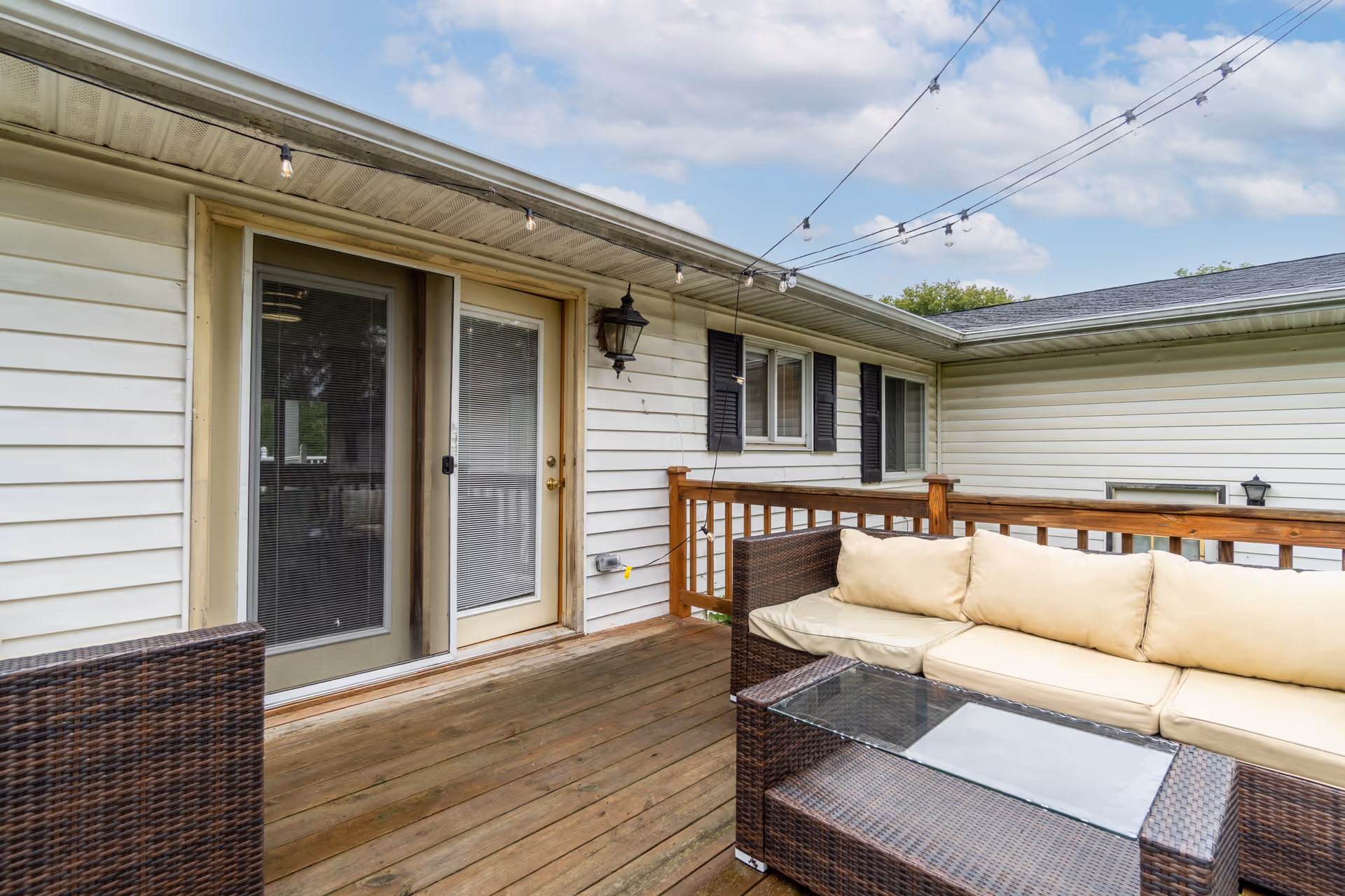 A wooden outdoor deck with wicker furniture and string lights beside a beige house.