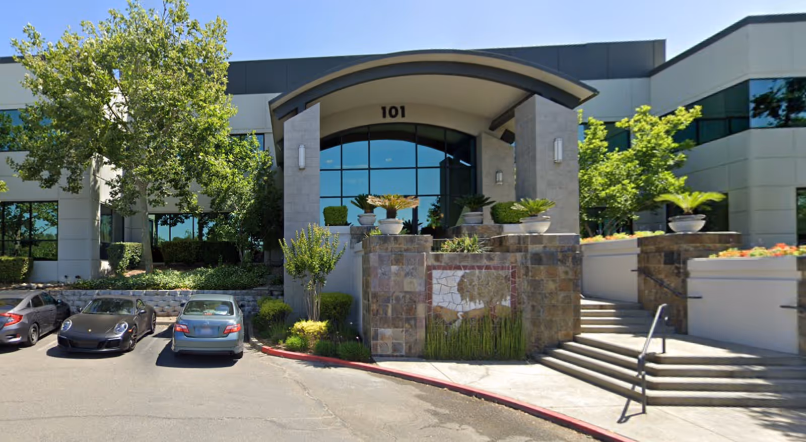 Building entrance with curved arch, steps, stone planters, and several parked cars.