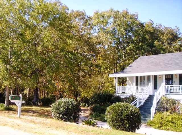 Single-story house with a front porch, steps, shrubs, and surrounding trees.