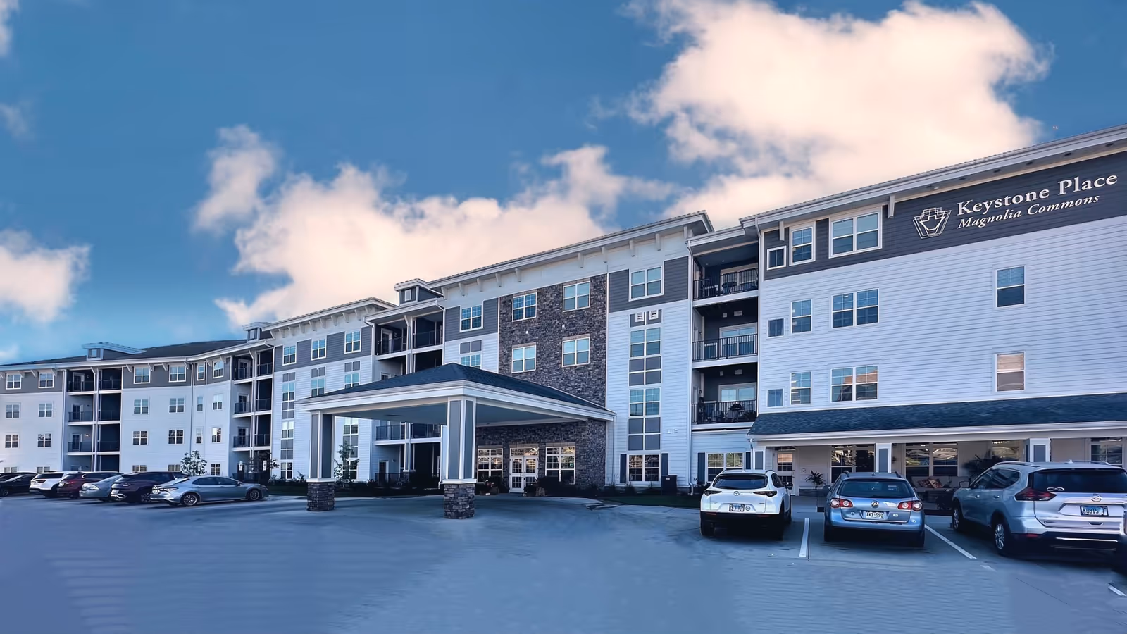 Front exterior of a multi-story senior living building with a covered porte-cochère, parked cars, and a sign reading "Keystone Place Magnolia Commons."
