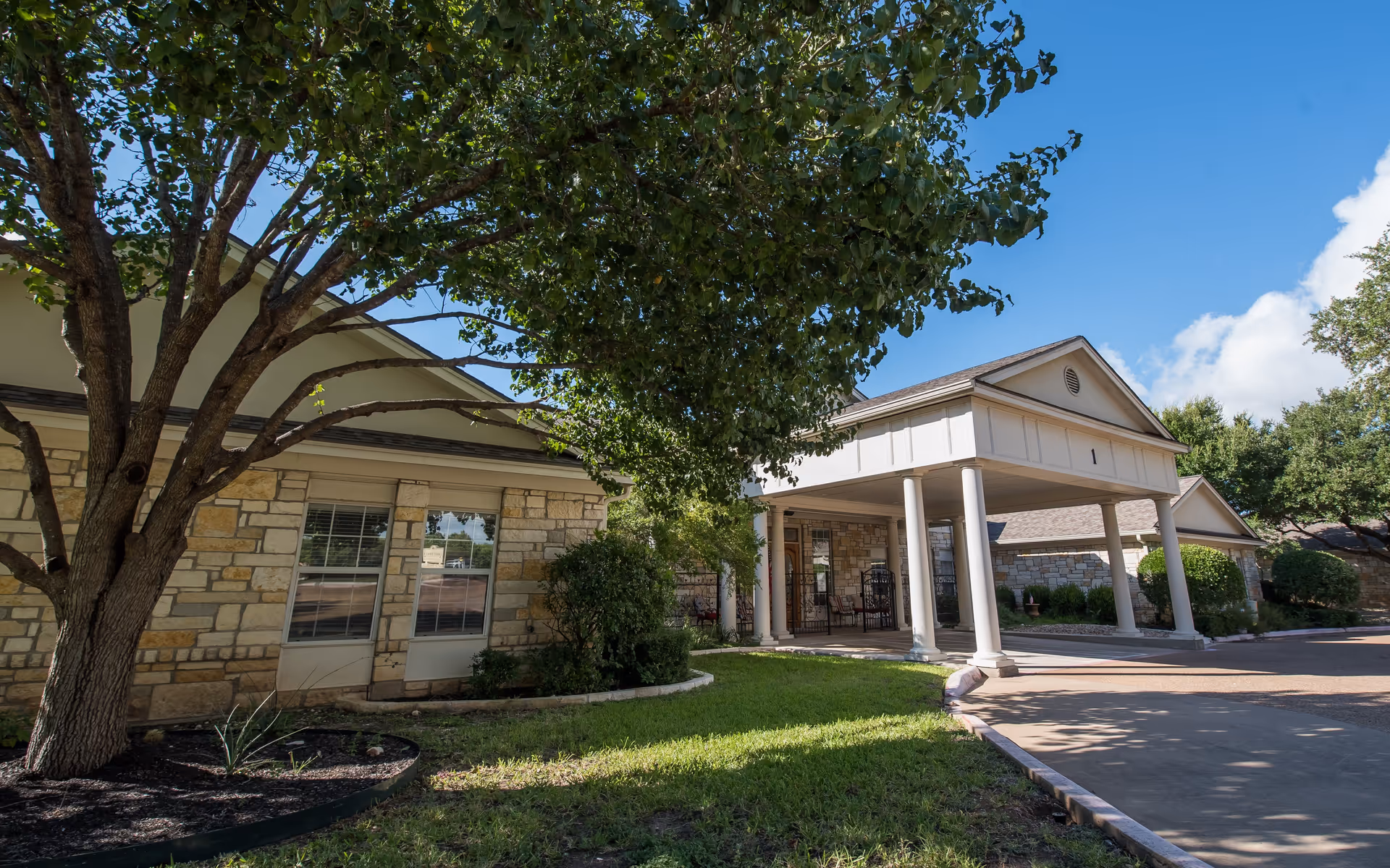 Exterior view of S.P.J.S.T. Assisted Living at Lake Travis showing a stone building with a covered entrance supported by white columns, surrounded by green trees and grass under a clear blue sky.