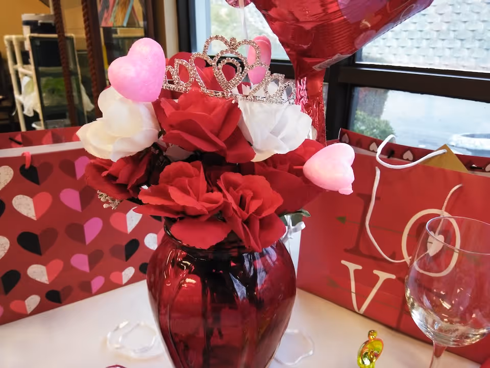Valentine's Day table centerpiece with a red vase of red and white roses, heart decorations, a small tiara, gift bags and a wine glass.