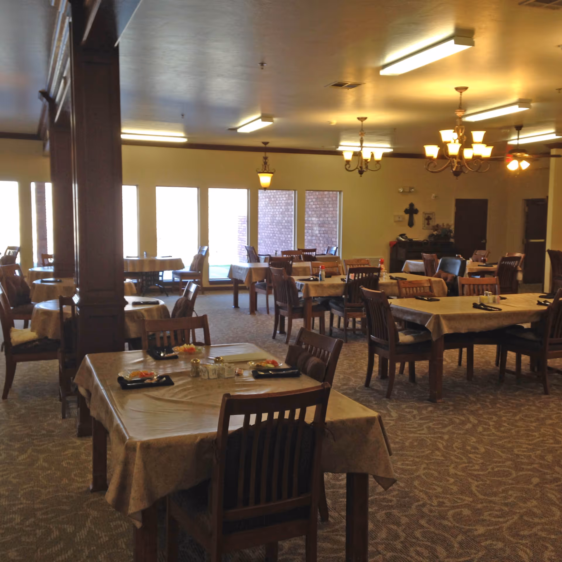 A spacious dining room with multiple tables covered in beige tablecloths, each set with black napkins and condiments. Wooden chairs surround the tables. The room is softly lit by ceiling lights and chandeliers, with large windows along one wall allowing natural light to enter.
