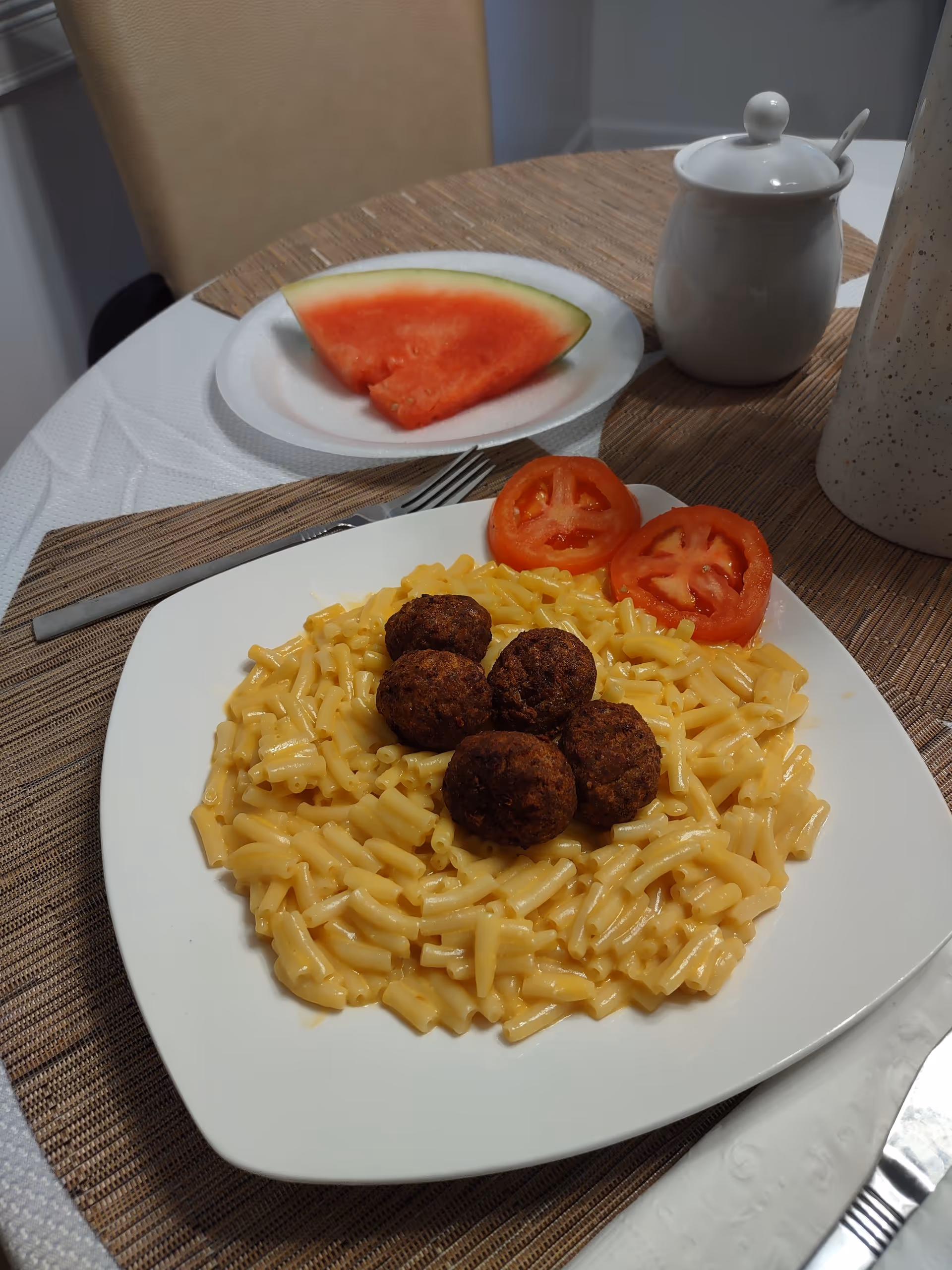Plate of pasta with meatballs on a dining table, watermelon slice nearby.