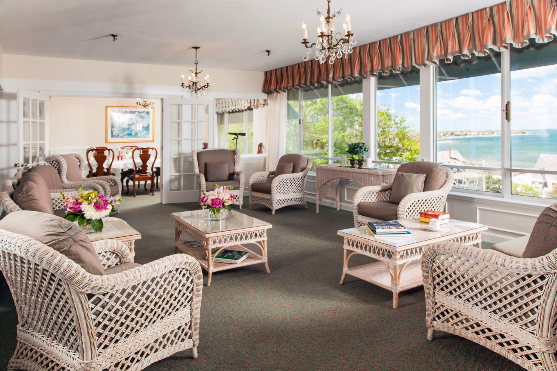 Bright lounge with wicker armchairs, coffee tables, and floral arrangements facing large windows with a water view.
