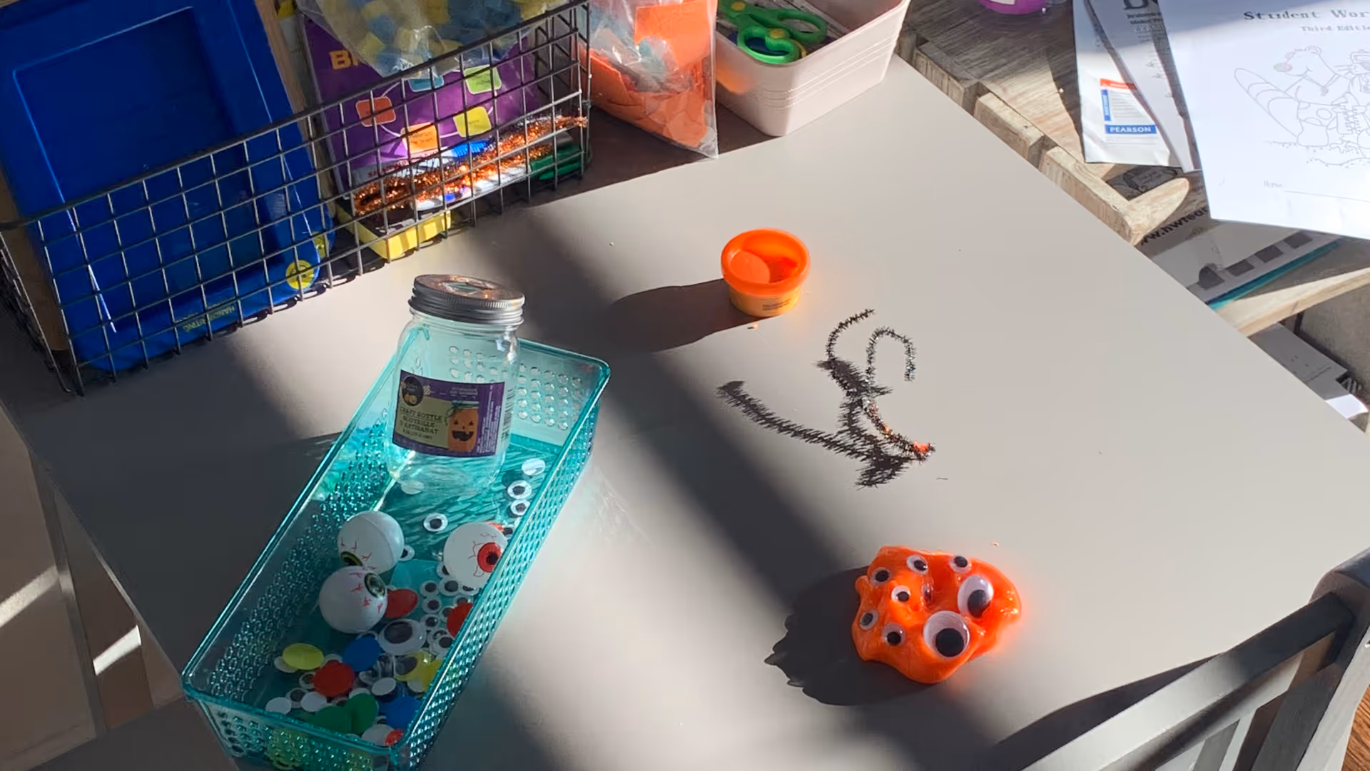 An indoor activity table with craft supplies, jars, and a small orange toy with googly eyes.