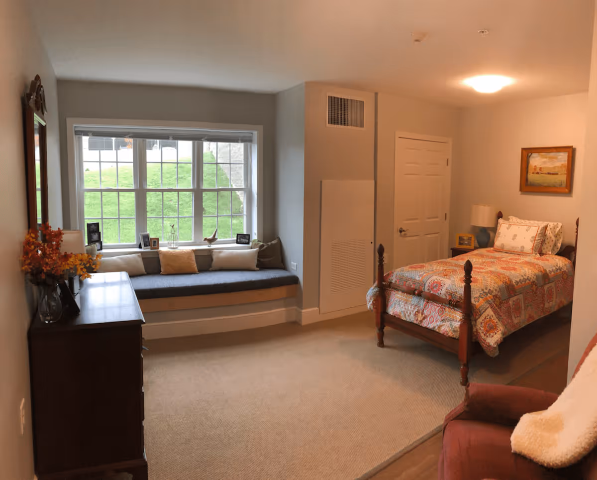 A cozy bedroom with a single wooden bed covered in a colorful quilt. There is a window seat with cushions beneath a large window showing green grass outside. A wooden dresser with a vase of flowers and framed photos is on the left side. A red armchair with a white throw blanket is partially visible in the foreground. The room has beige carpet and soft lighting from a ceiling fixture and a bedside lamp.