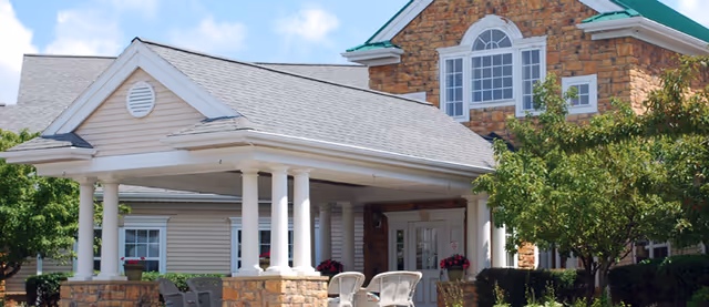 Covered entrance of a senior living building with white columns, stone and siding facade, outdoor chairs and landscaping.
