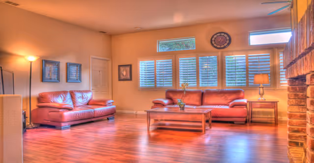 A spacious living room with two red leather sofas, a wooden coffee table with a small plant, a side table with a lamp, framed pictures on the walls, large windows with white shutters, and a brick fireplace on the right side.
