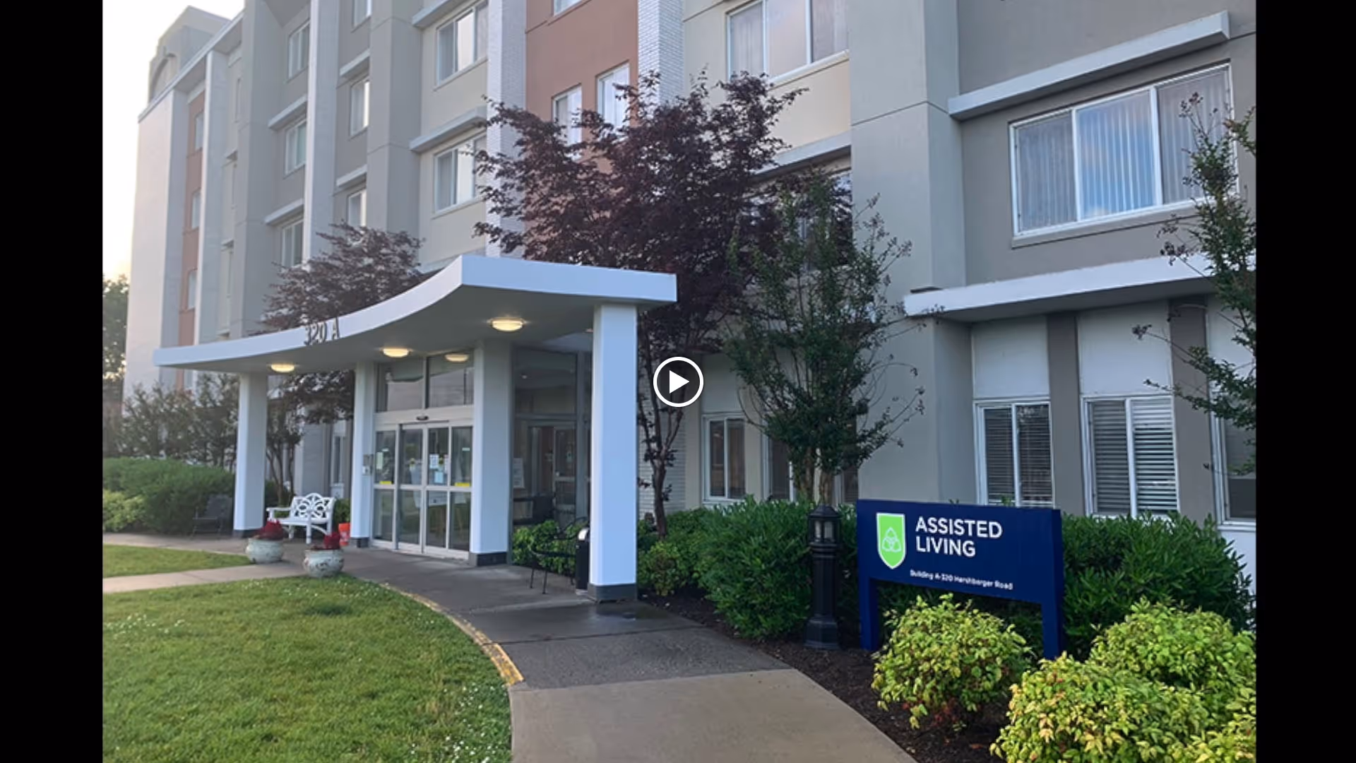 Exterior view of a multi-story assisted living facility building with a covered entrance, surrounded by greenery and trees. A blue sign near the entrance reads 'ASSISTED LIVING Building 4 320 Norrlinger Road'.