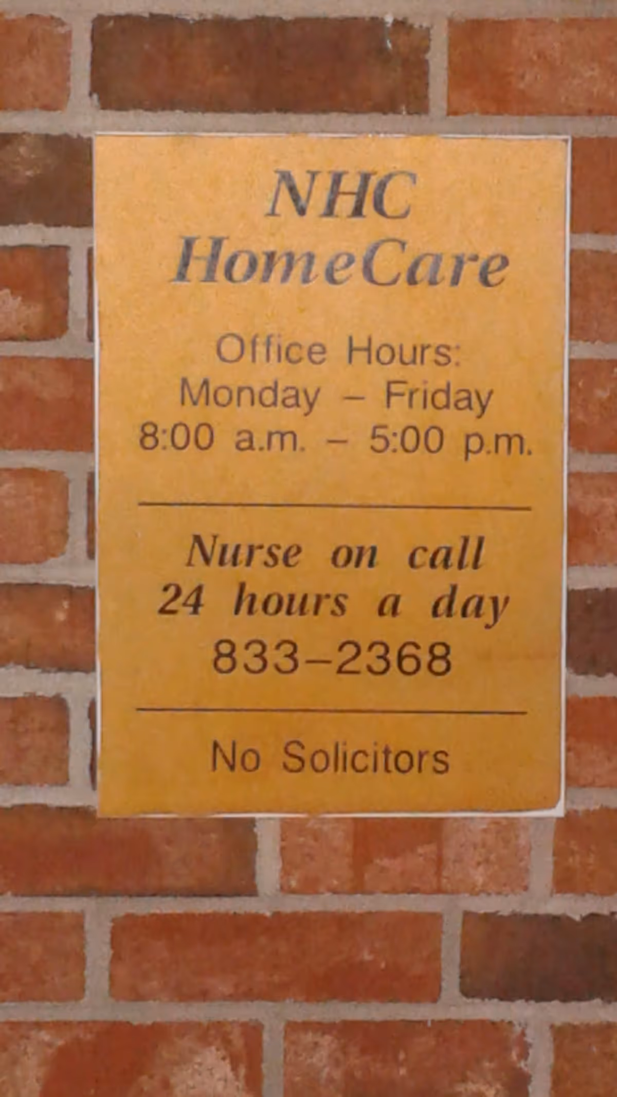 Yellow sign on brick wall with NHC HomeCare office hours and phone number.