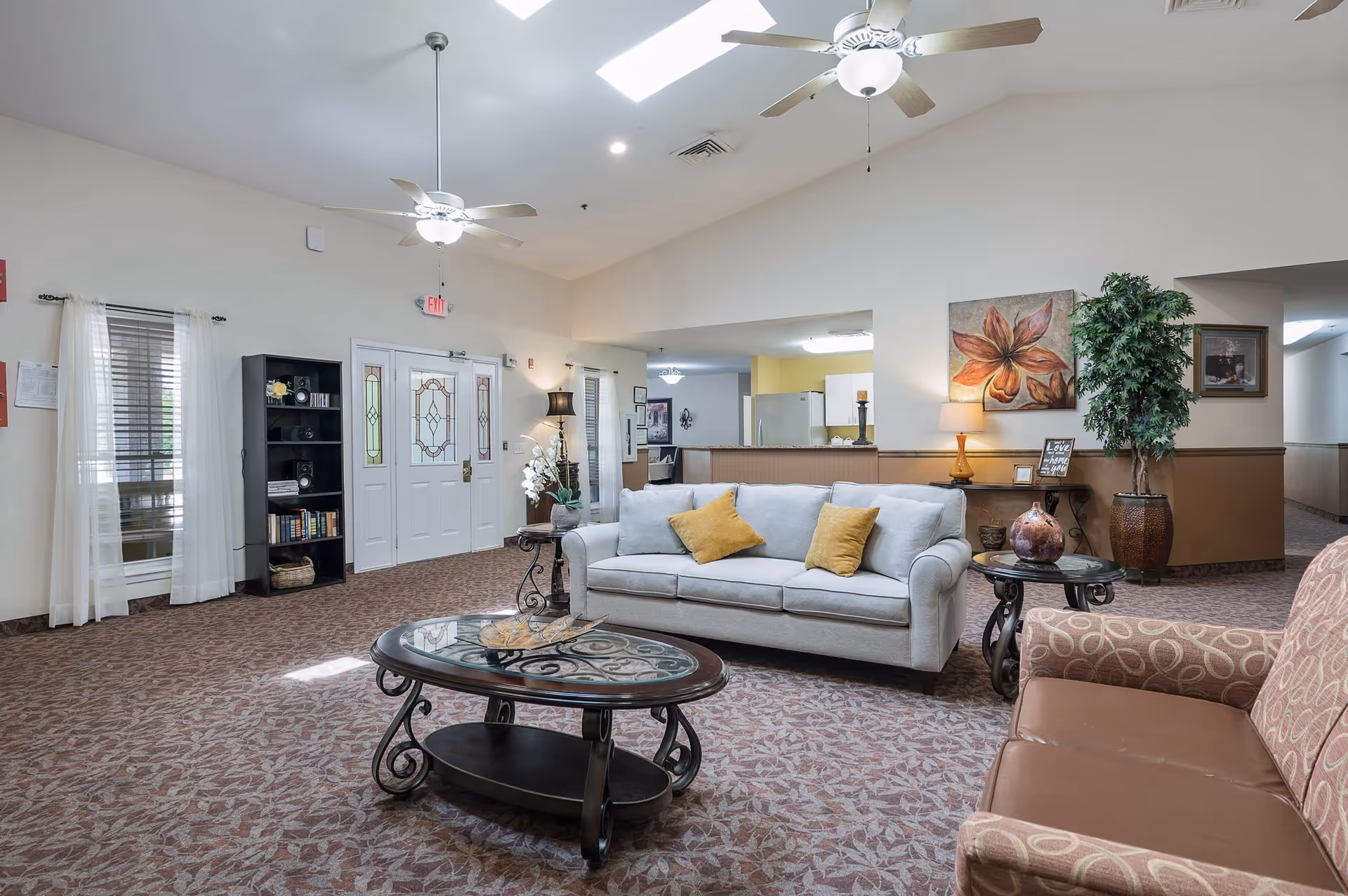 A spacious and well-lit living room area in an assisted living facility featuring a white sofa with yellow cushions, a patterned armchair, a glass-top coffee table, and a side table with a decorative vase. The room has carpeted floors, ceiling fans with lights, a bookshelf, and a large painting of a flower on the wall. There is a view into a kitchen area with white cabinets and a refrigerator.