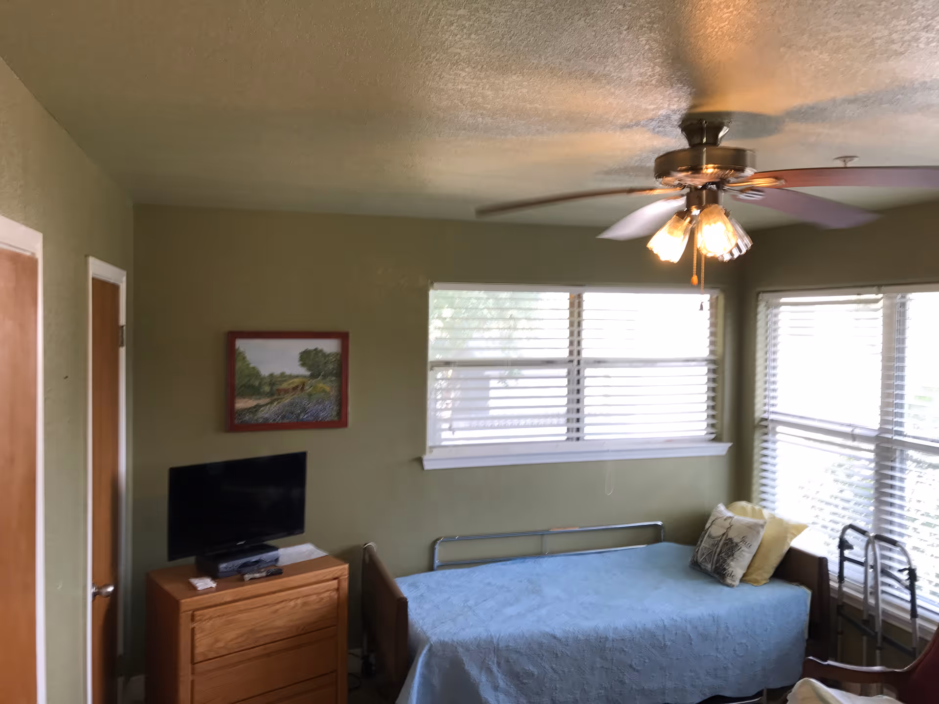 Green-walled bedroom with a blue bed, dresser, small TV, and window blinds.