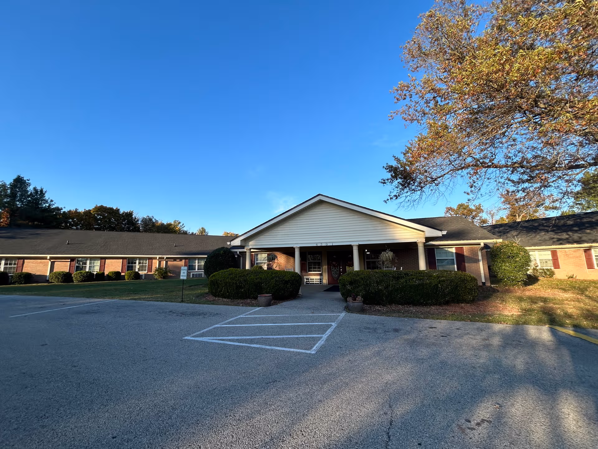 Single-story brick senior living facility with front entrance and manicured shrubbery under a blue sky.