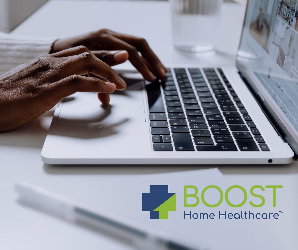 Close-up of hands typing on a laptop at a desk displaying the BOOST Home Healthcare logo.