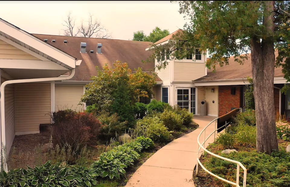 A paved walkway with a handrail winds through landscaped gardens to the front entrance of a single-story senior living building.