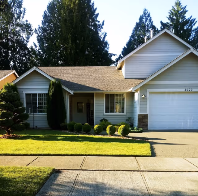 Exterior front view of a single-story residential house with a well-maintained lawn, shrubs, and tall trees in the background. The house has light-colored siding, a two-car garage, and a concrete driveway and sidewalk in front.